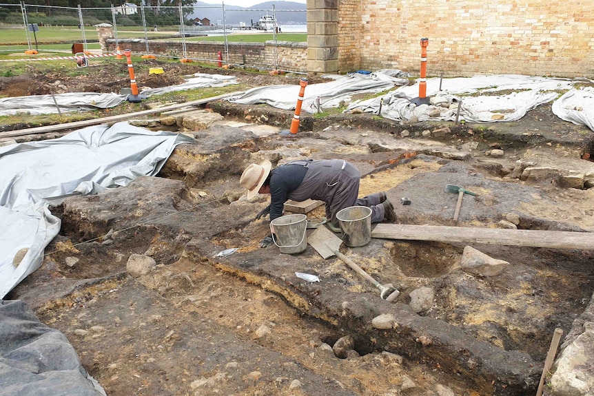 An archaeological dig with a woman wearing overalls and a hat on her knees working on brown dirt