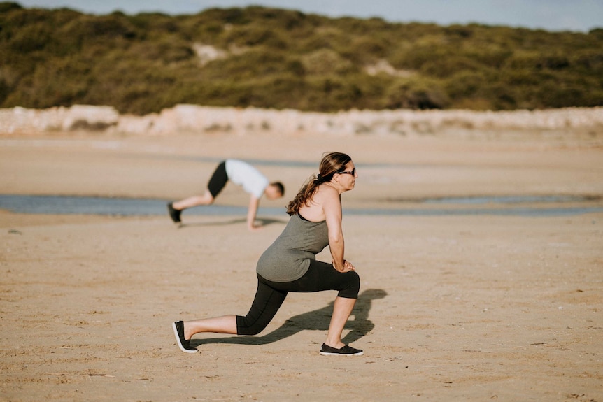 Woman lunging on beach with man in the background to depict how to stick to fitness goals and daily exercise habits.