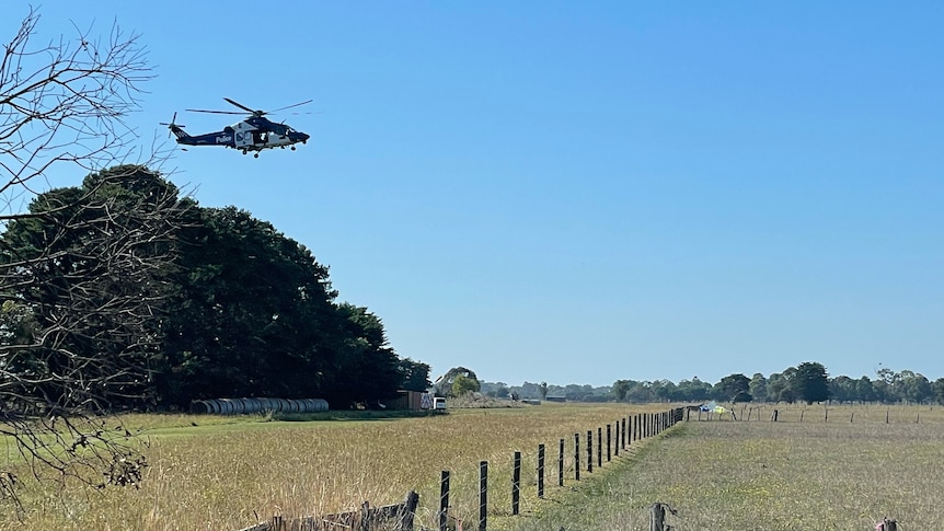 A helicopter flies overhead as emergency services inspect the scene of a light plane crash in the distance.