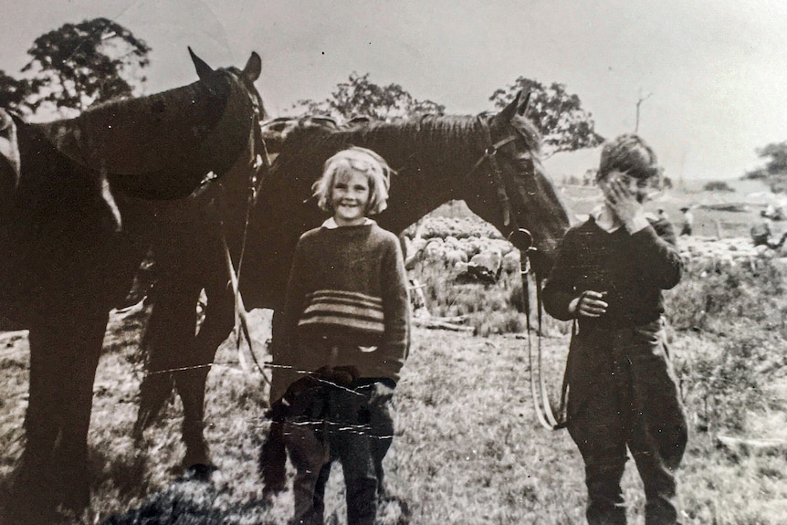 A black and white photo of a small girl and boy with their horses and a mob of sheep in the background.