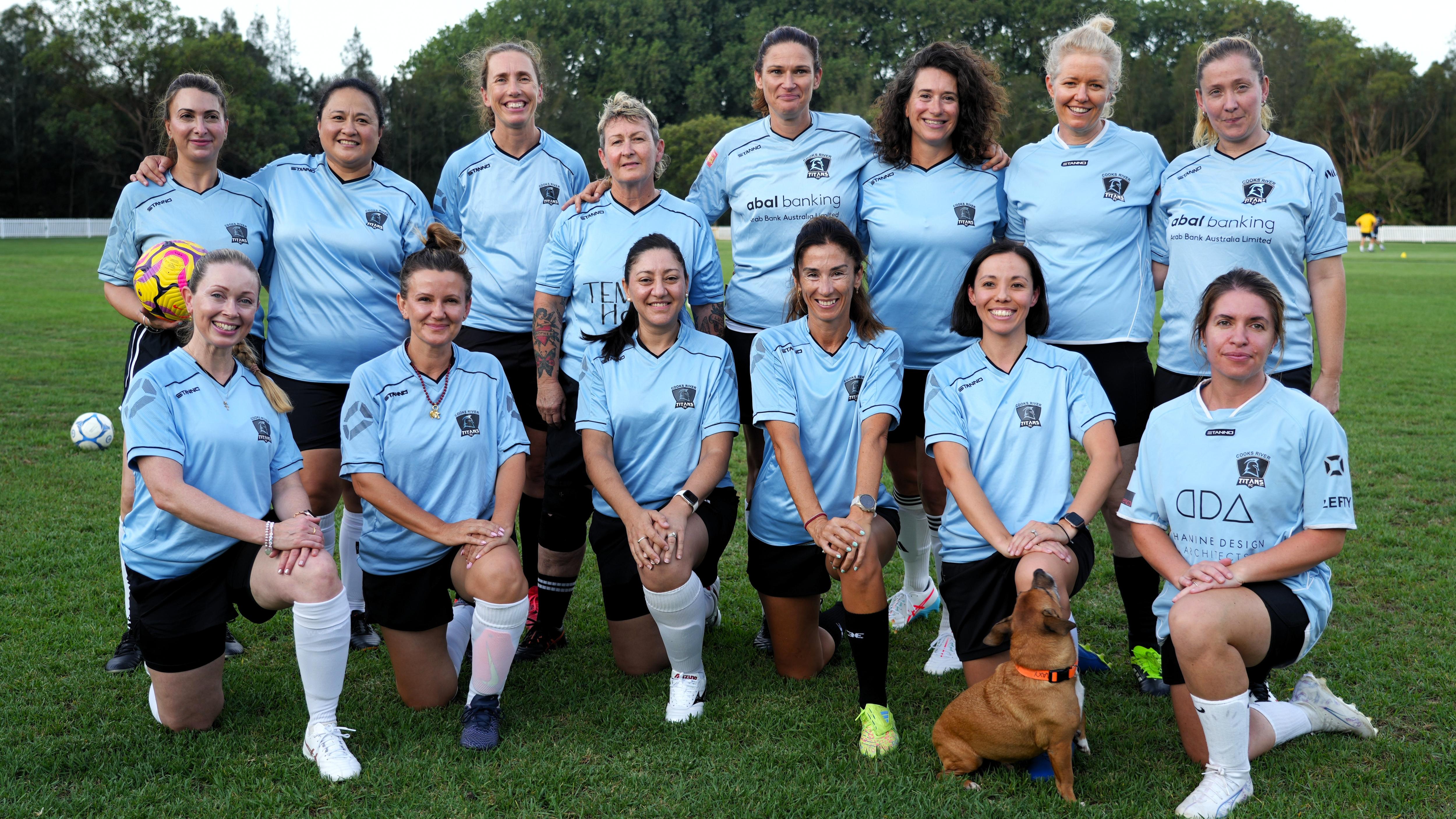 Membros de um time de futebol feminino com mais de 25 anos se alinham em duas fileiras e sorriem para uma foto do time.