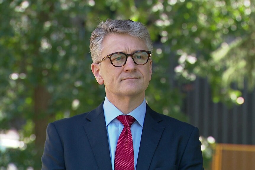 Chris Blake wears a blue jacket, light blue collared shirt, red tie and brown rimmed glasses and stands outside.