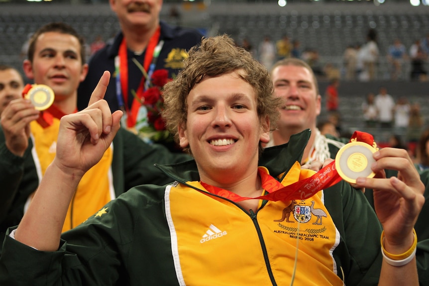 Wheelchair basketball player Dylan Alcott holds his Paralympic gold medal in one hand and points up with his other hand.