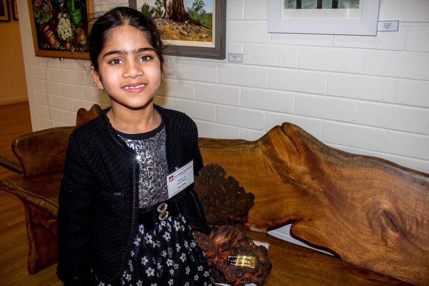 Esha Jabbal stands in front of a bench indoors holding a Dorothea Mackellar Poetry Award and smiling for the camera.