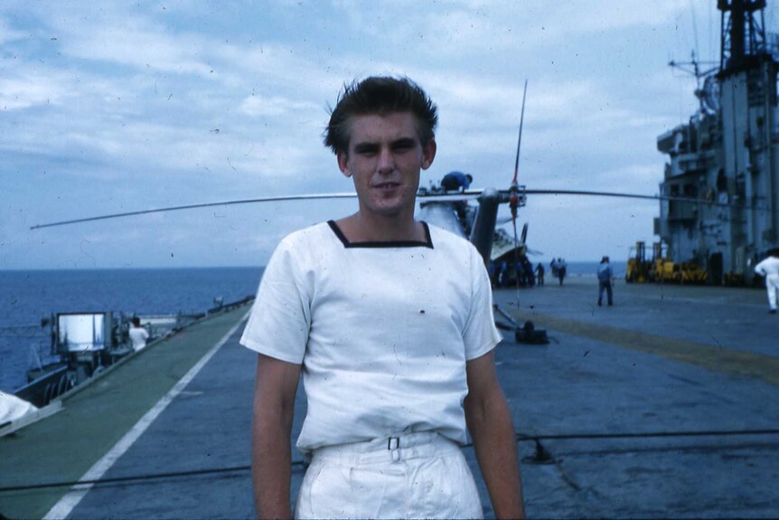 Young Navy man in whites standing on deck of aircraft carrier with helicopter in background on deck 