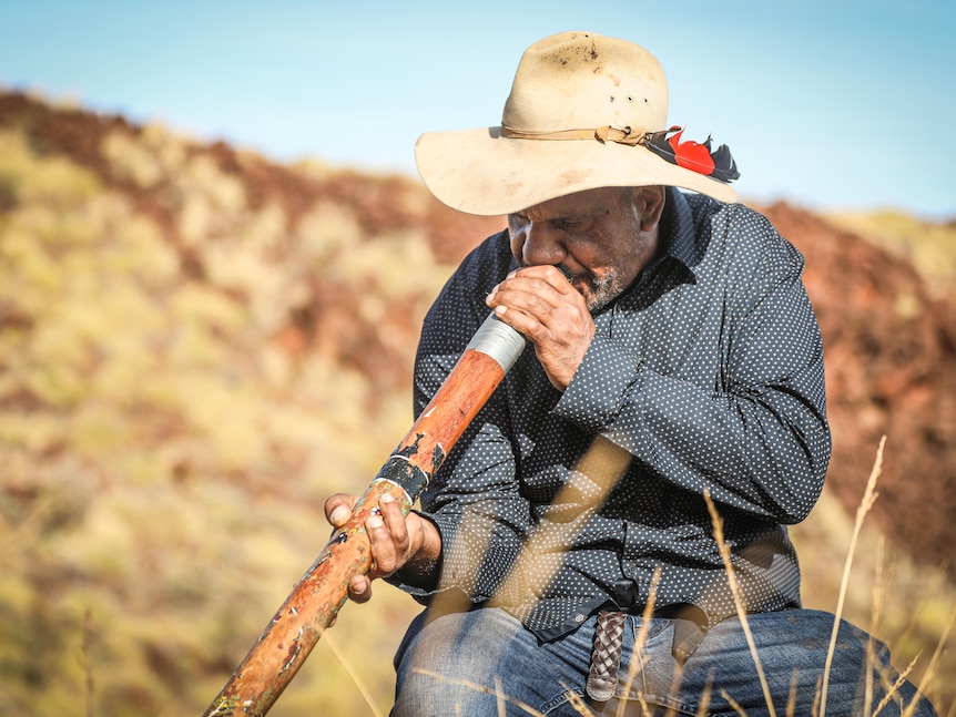Image of an older man wearing an akubra, sitting down and playing the didgeridoo