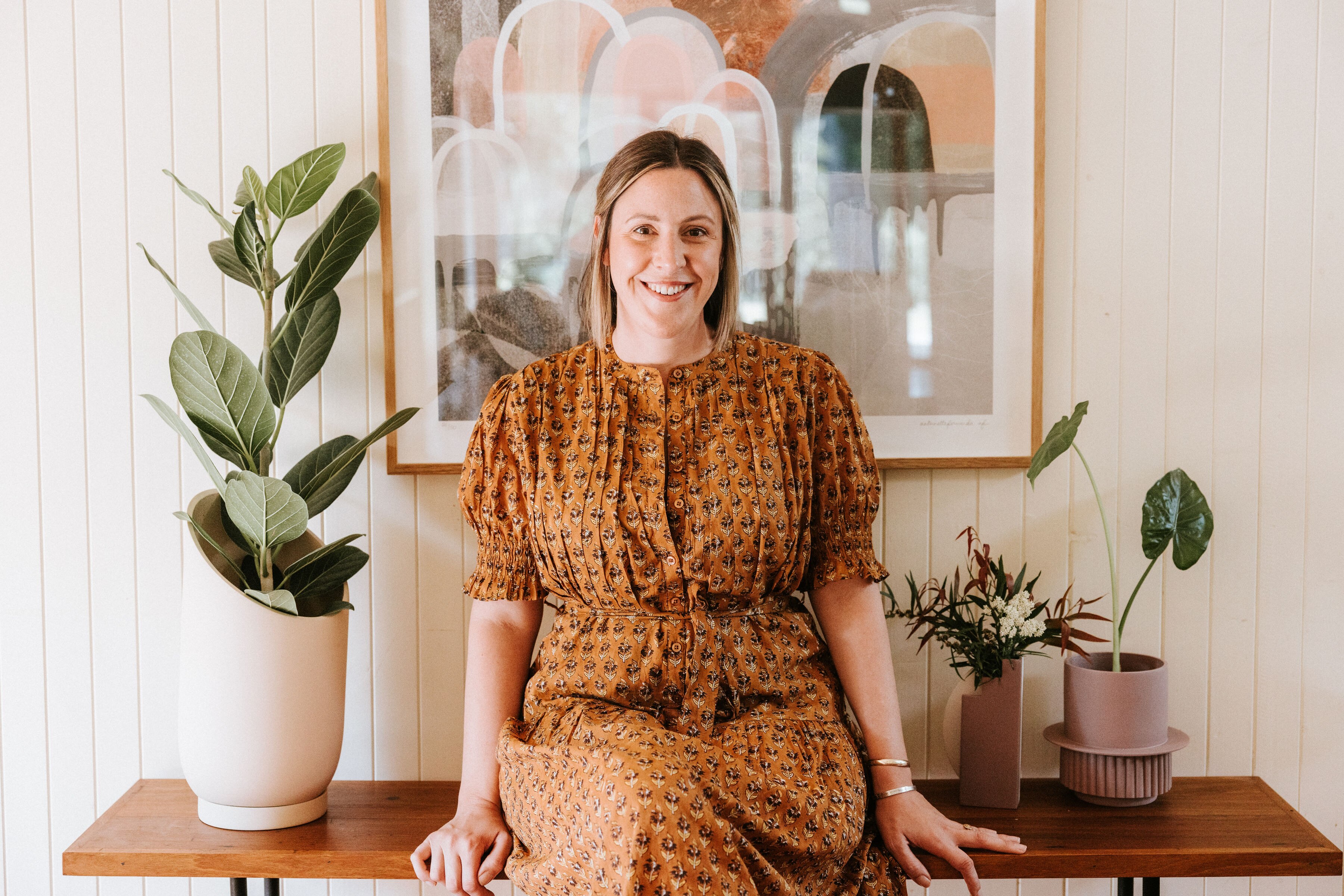 A woman stands in front of a desk with pot plants on it, and a painting hangs on the wall.