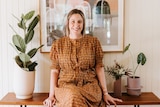 A woman stands in front of a desk with pot plants on it, and a painting hangs on the wall.