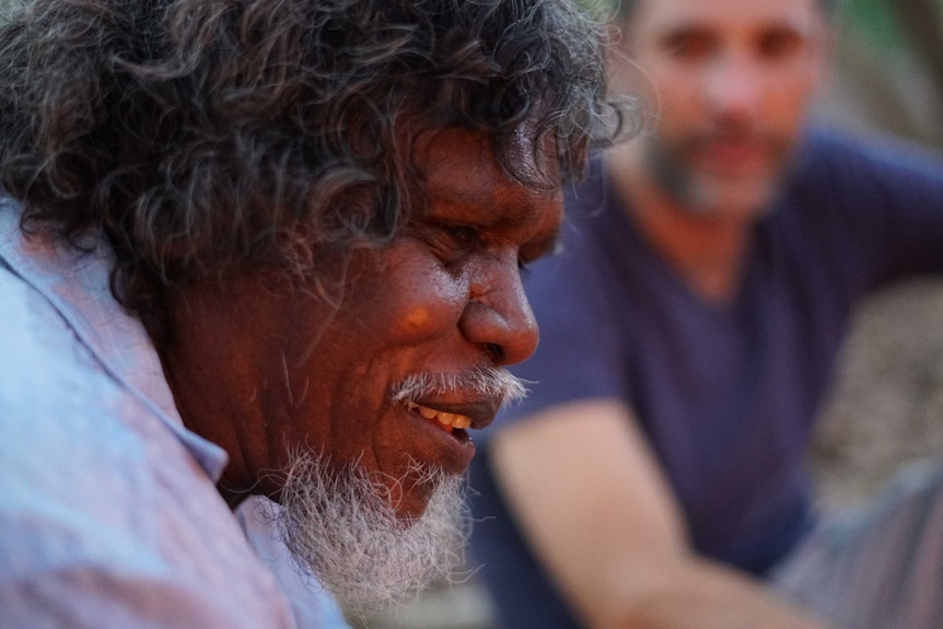An Indigenous man is smiling in the foreground with another man blurred in the background.