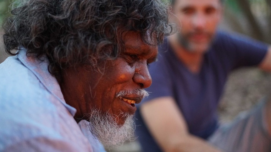 An Indigenous man is smiling in the foreground with another man blurred in the background.