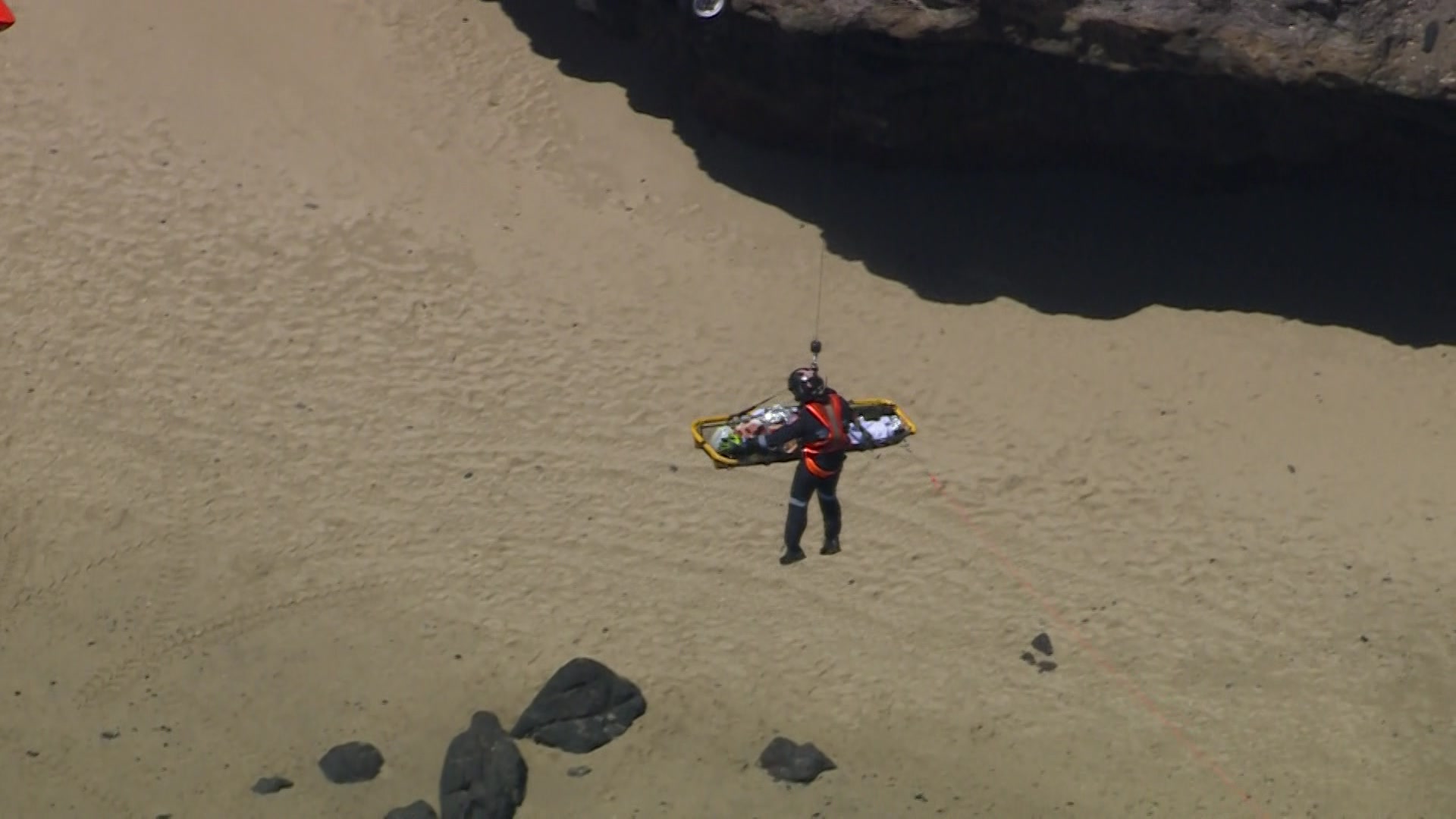 Una persona que lleva un casco oscuro, un mono y un arnés naranja cuelga en el aire con una persona en una camilla sobre rocas y agua.