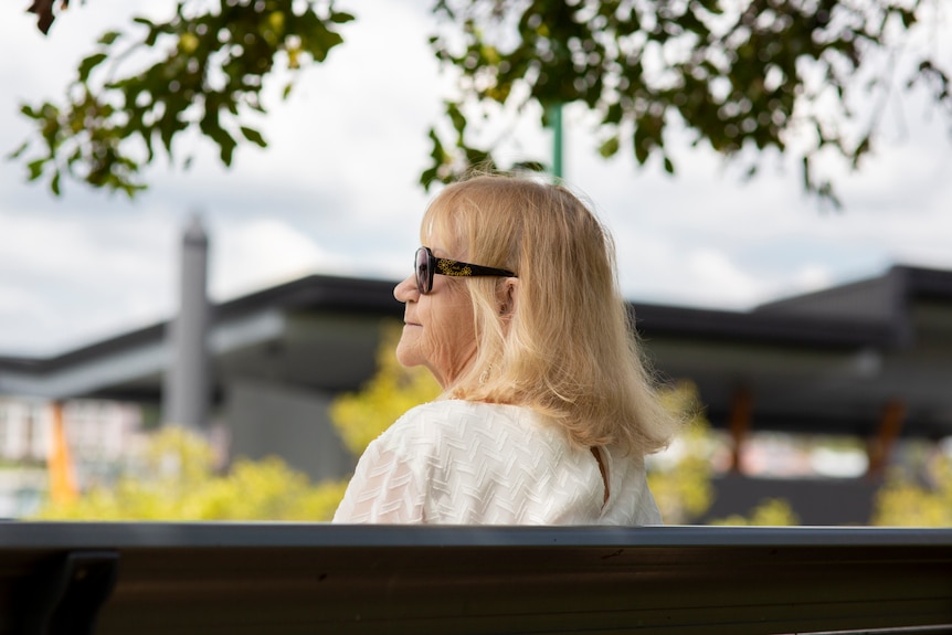 A woman with a blonde bob and dark sunglasses looks to her left and stares into the distance.