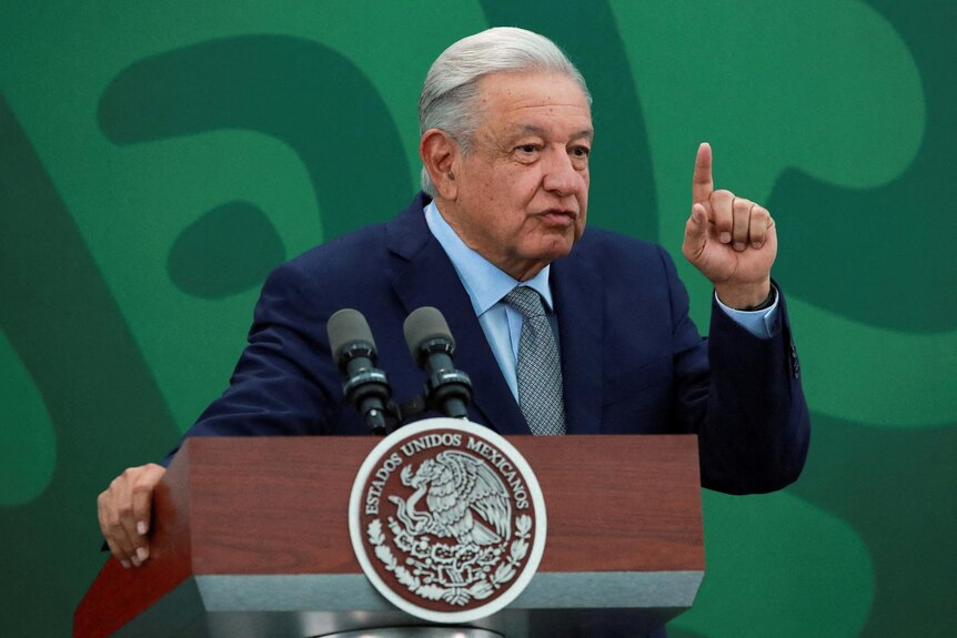 An older Mexican man in a blue suit points as he speaks behind a lectern.
