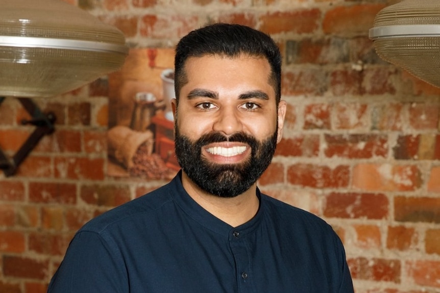 Man with beard and black shirt smiles to camera in front of brick wall. 