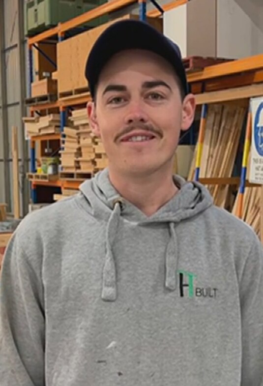 A young man with a moustache wears a dark cap and grey hoodie in a construction warehouse