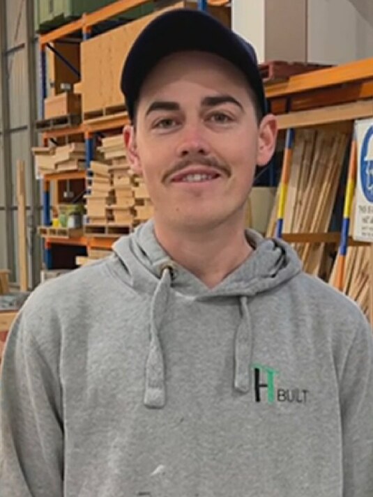 A young man with a moustache wears a dark cap and grey hoodie in a construction warehouse
