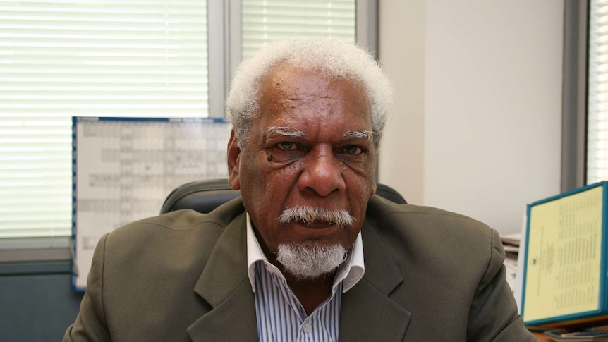 An Aboriginal man with grey hair wearing a beige suit sitting in his office.