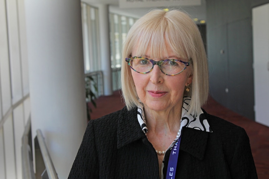 Mature-aged lady with colourful glasses and blonde hair at a conference.