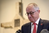 Prime Minister Malcolm Turnbull stands at a microphone looking down with a cross behind him.