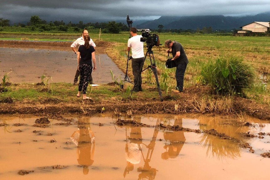 Cameraman, reporter, producer and sound recordist filming in flooded field with dark clouds overhead.