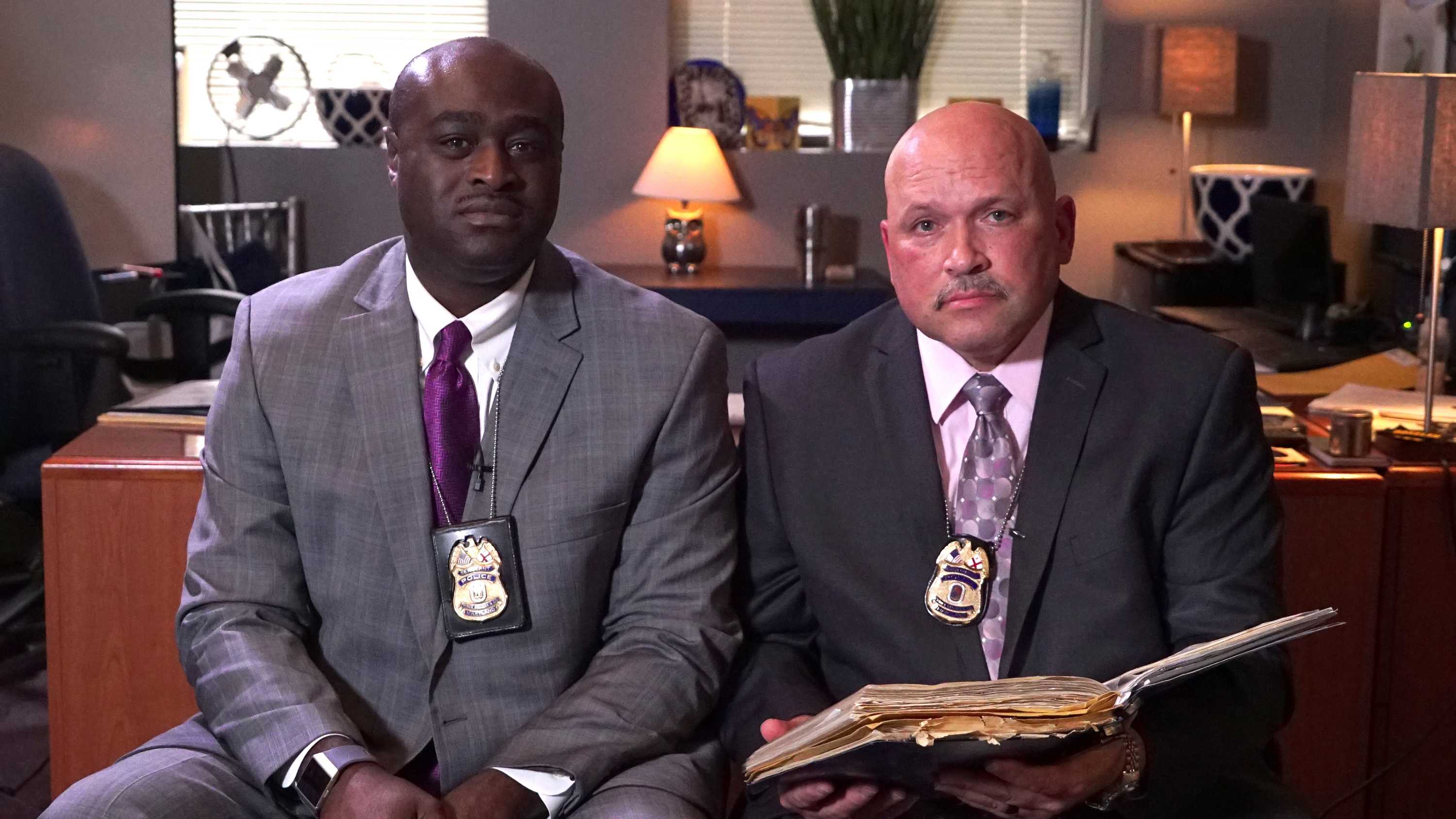 African-American police officer Gregory McDonald sits down next to white police officer Bernard Nelson with a desk behind them.