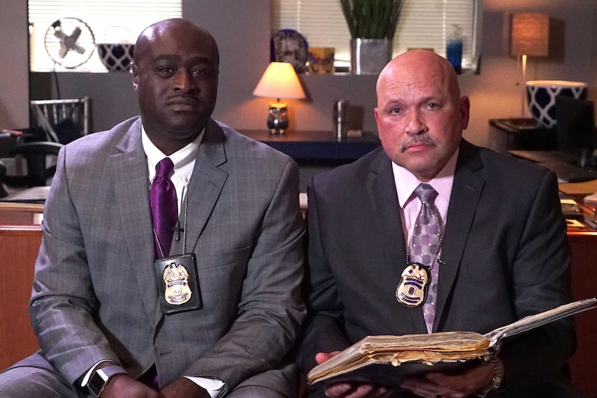 African-American police officer Gregory McDonald sits down next to white police officer Bernard Nelson with a desk behind them.