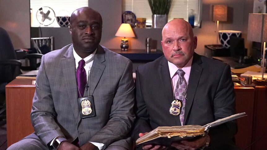 African-American police officer Gregory McDonald sits down next to white police officer Bernard Nelson with a desk behind them.