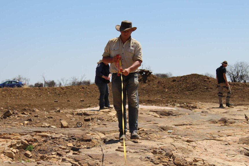 A man wearing dusty clothes and a broad-brimmed hat unspools a yellow tape along rocky ground.