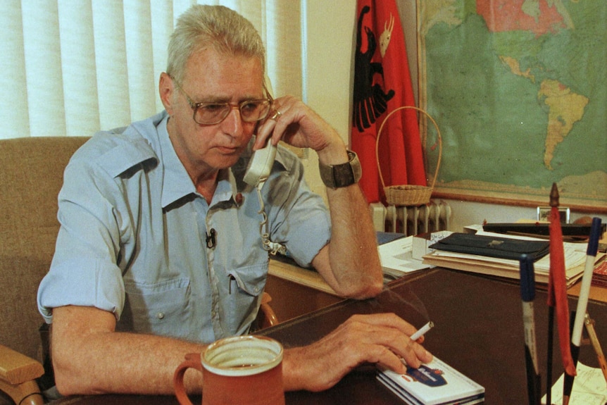King Leka, a middle-aged man with white hair, sits at a desk holding a phone to his ear and cigarette in the hand