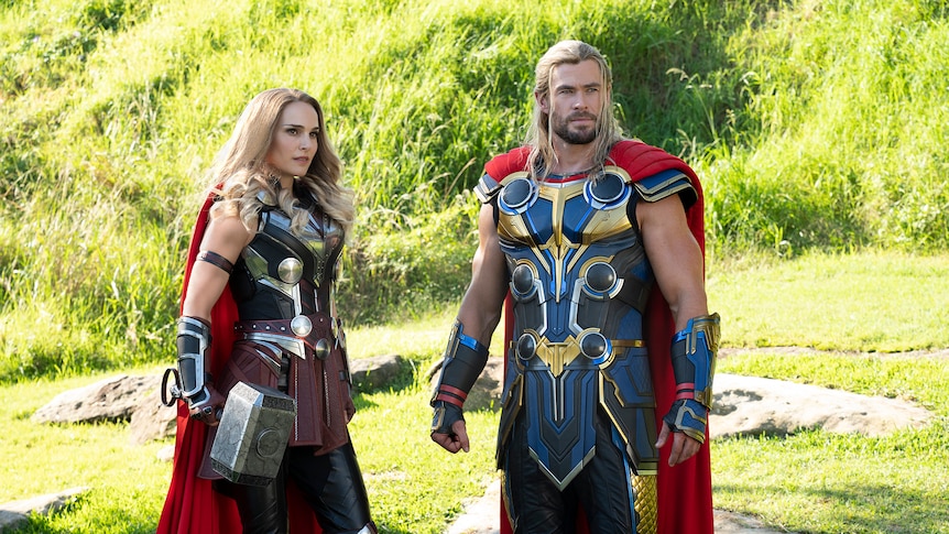 White woman with curled blonde hair stands with man with long blonde hair. They both wear blue and gold armor and a red cape.