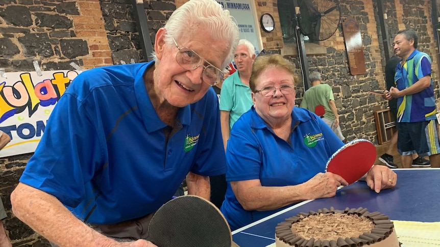 Elderly couple hold table tennis bats and smile after being presented with a chocolate cake