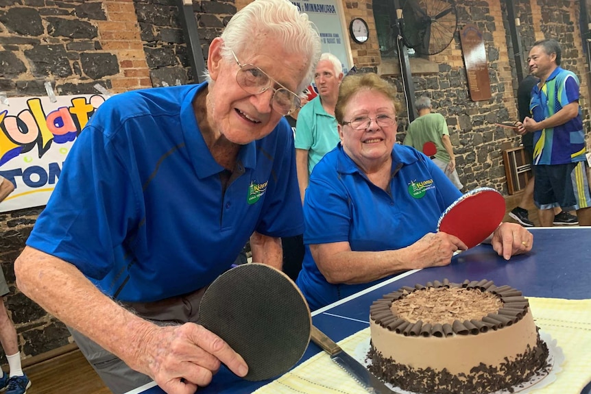 Elderly couple hold table tennis bats and smile after being presented with a chocolate cake