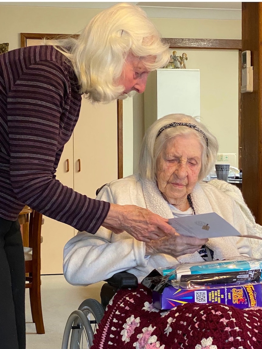 An elderly woman sits in a wheelchair with a maroon blanket over her lap, while another white-haired woman hands her a card.