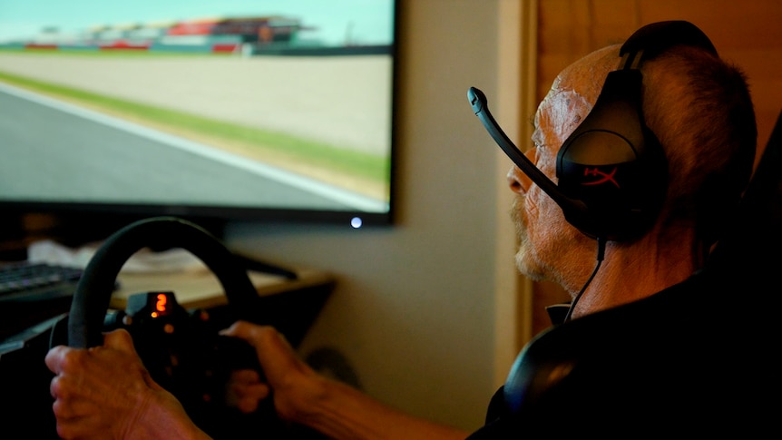 An old man wearing a gaming headset, holding a video game steering wheel.