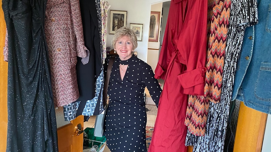 A smiling older woman in a stylish dark dress, standing among hanging clothes.