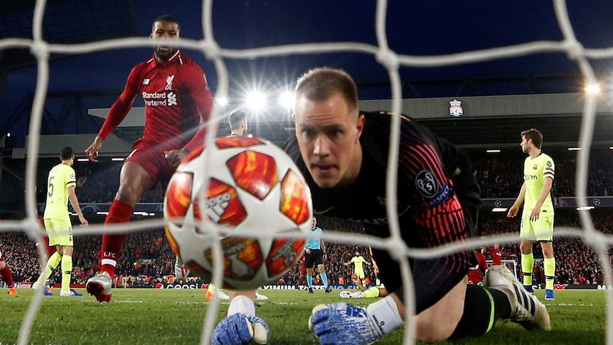 A goalkeeper kneels down and watches the ball in his net as opposing players celebrate a goal under flood lights