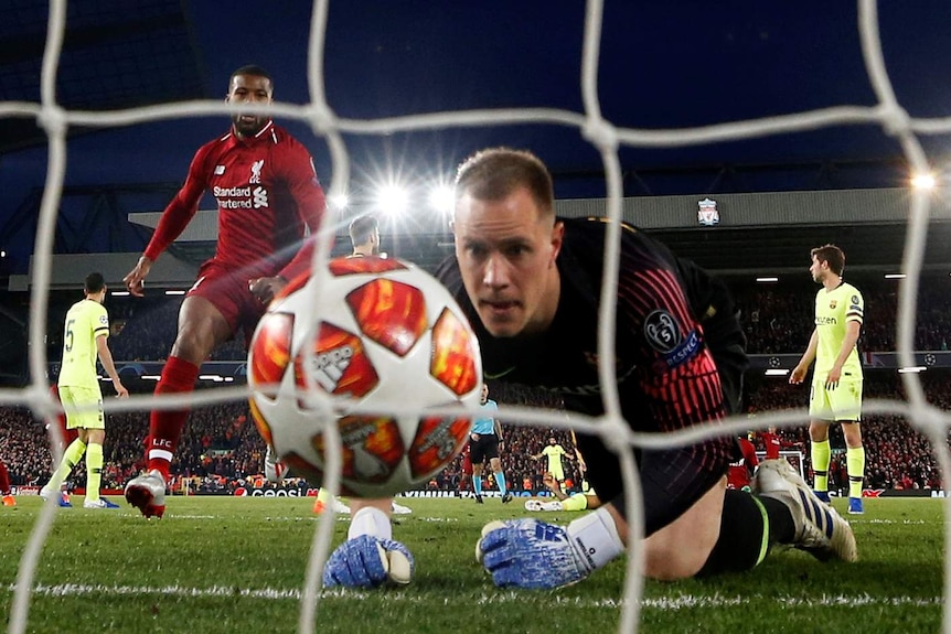 A goalkeeper kneels down and watches the ball in his net as opposing players celebrate a goal under flood lights