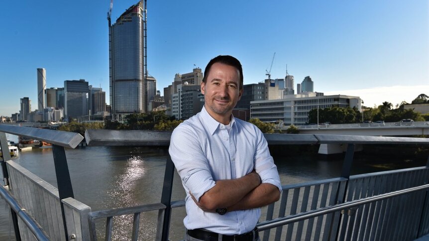 Marcus stands arms folded on a Brisbane bridge with the CBD in the background.