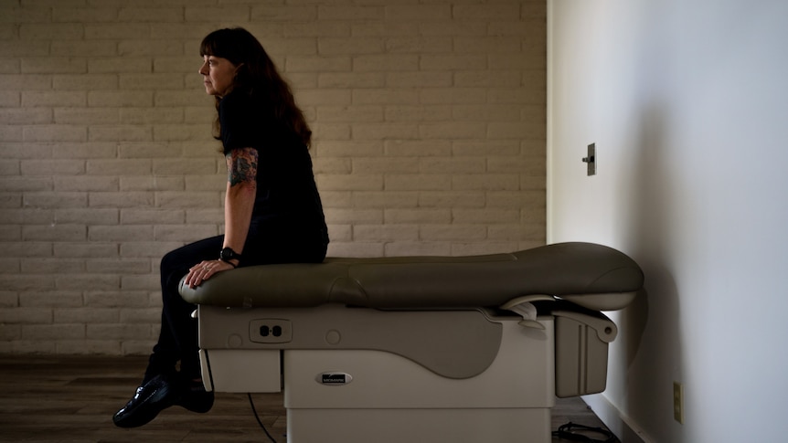 A woman with a tattoo sleeve sits on a hospital bed in an otherwise empty room