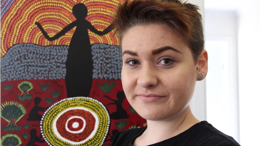 A young woman, Jamira Pemberton, standing in front of her painting of a dreamtime story