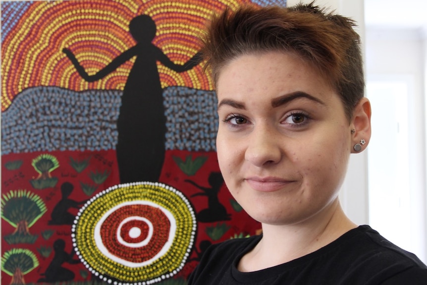 A young woman, Jamira Pemberton, standing in front of her painting of a dreamtime story