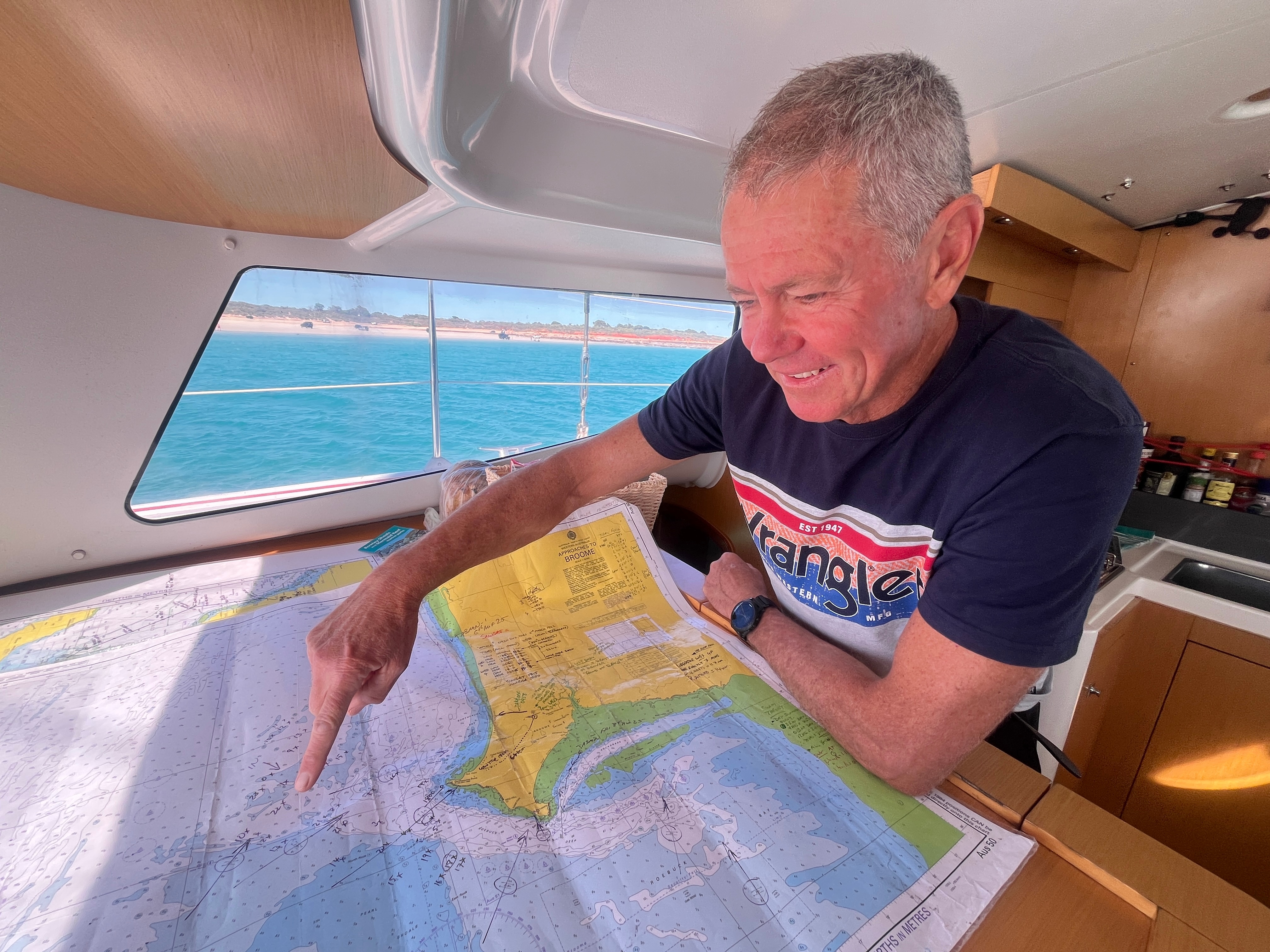 A man in a blue t-shirt points at a nautical map laid out in the cabin of a boat.