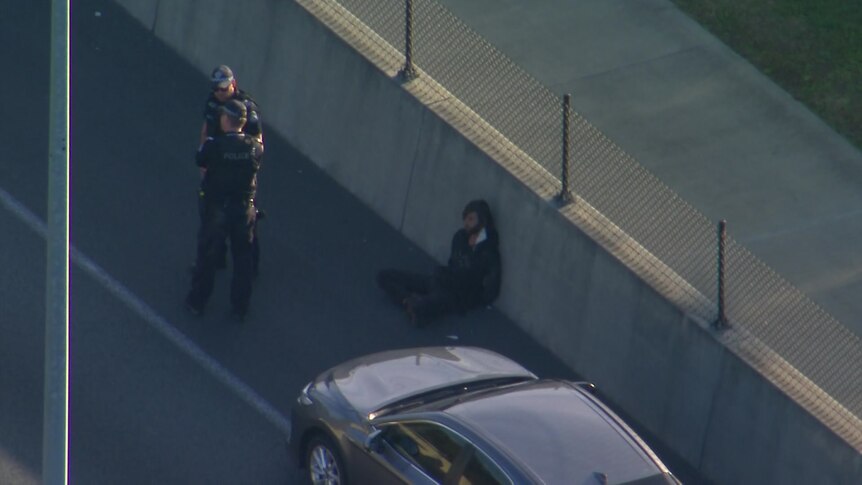 A person sits on the side of the road in handcuffs while two police officers stand nearby