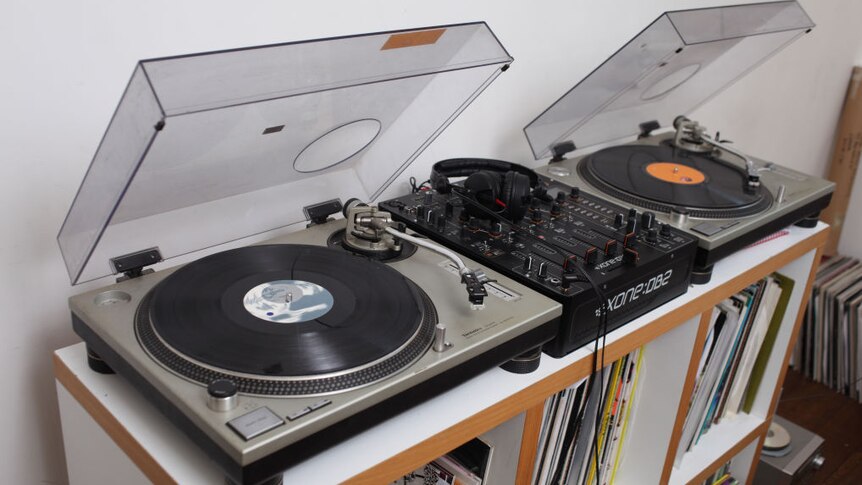 image shows 2 record players, a simple sound mixer and shelves of records.
