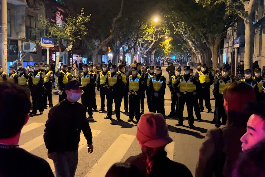 Chinese police officers stand in a line on a street blocking off protesters gathered on the other side of a pedestrian crossing.