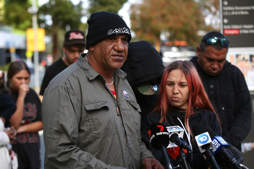 A tall man wearing a grey jacket and a black beanie stands in front of news microphones while a woman with pink hair stands.