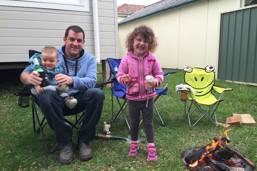 A man wearing a blue hoodie and jeans holds a happy baby boy in backyard. A toddler girl stands next to them