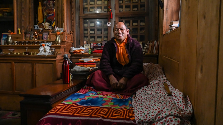 A man wearing monk's robes sits cross-legged on a bed covered in colourful fabric with a bookshelf and cabinet behind