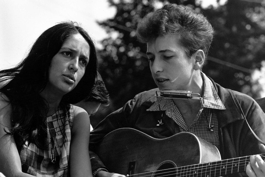 A young woman singing with a furrowed brow, next to a young man with a guitar and harmonica.