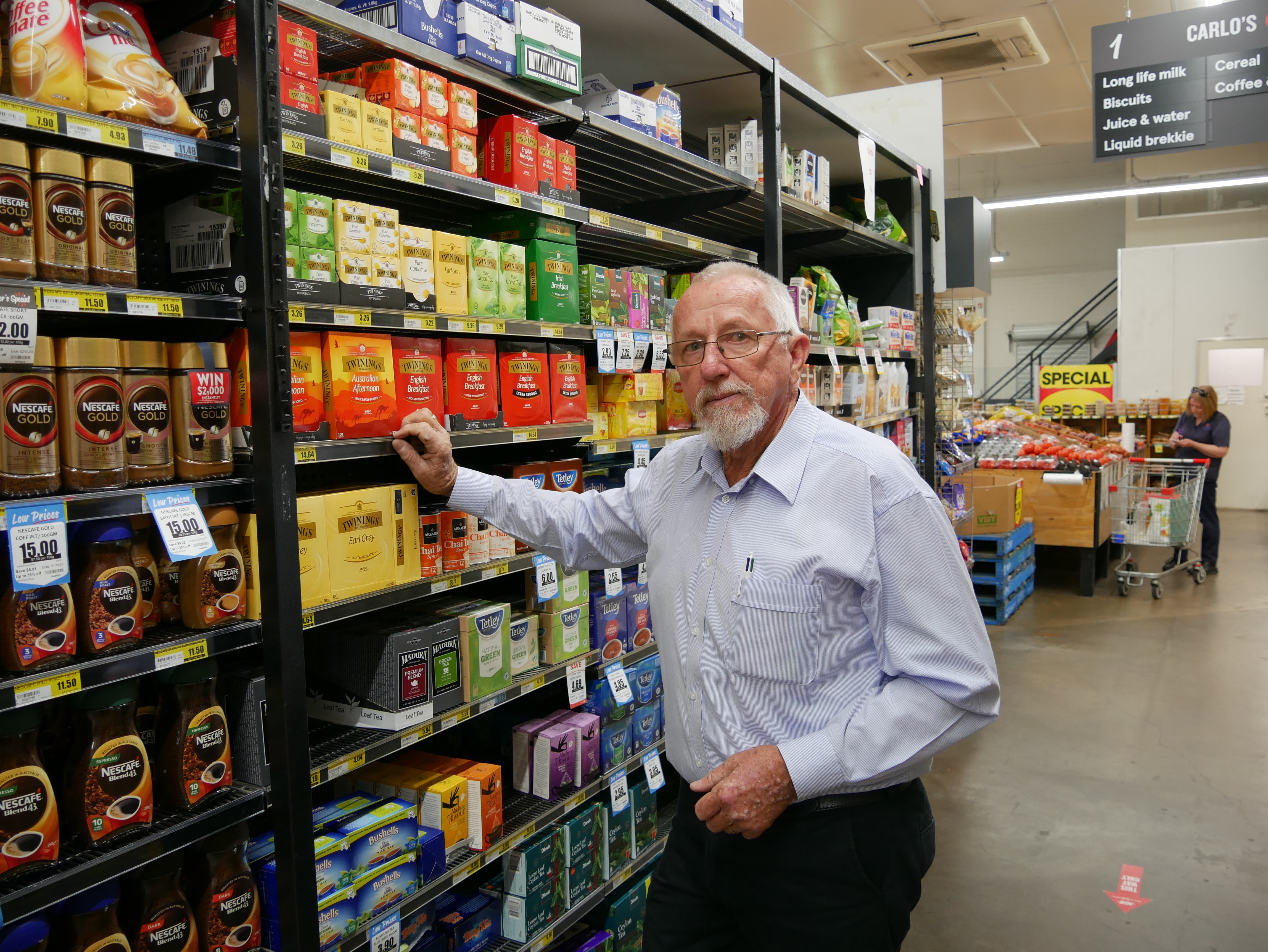 An elderly man with a blue button up shirt stands next to the grocery shelves in a retail store